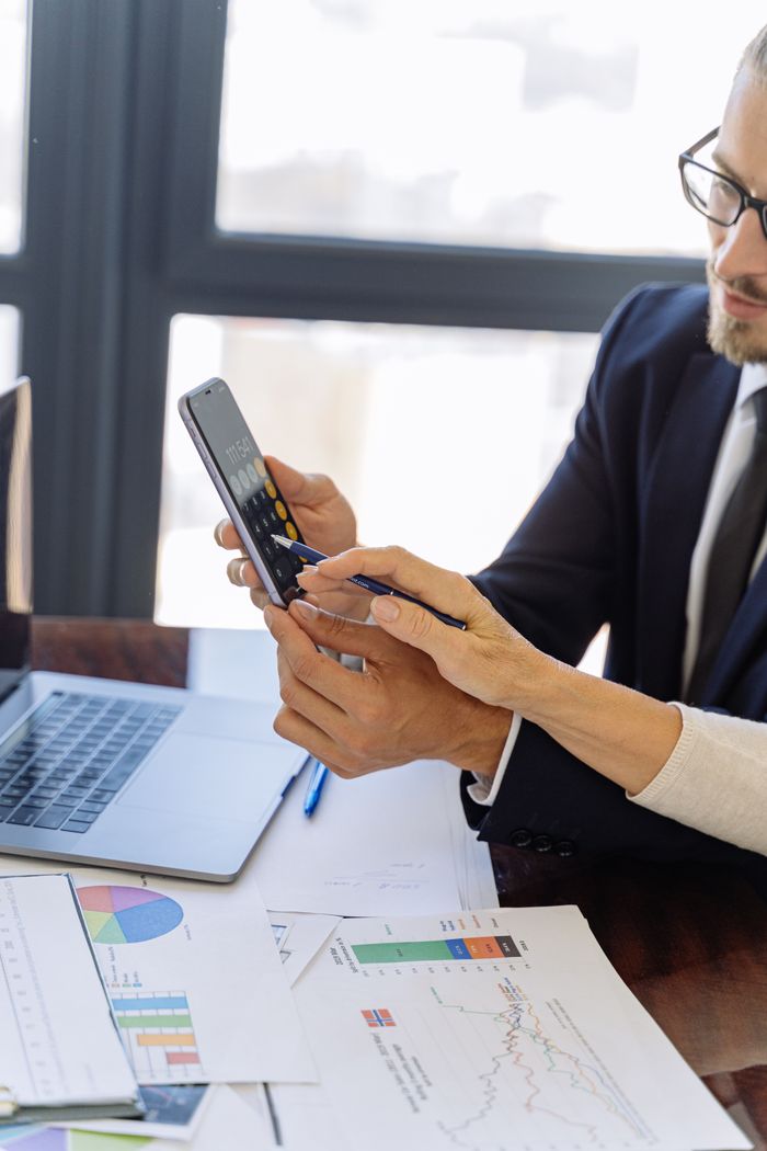 about-04 Business professionals reviewing financial reports on a smartphone at a desk with papers and a laptop in an office setting.